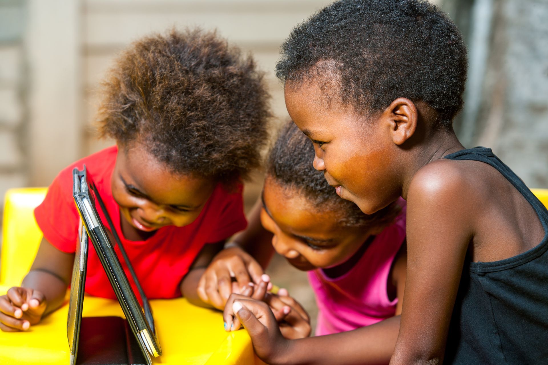 Three children leaning over a tablet, smiling and pointing together outdoors