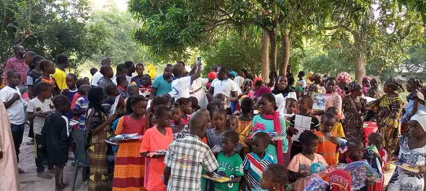 Crowd gathered outdoors under trees, with people seated and standing in colorful clothing