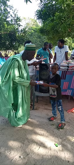 Three people outdoors sharing items, with a child standing beside them near chairs and trees.