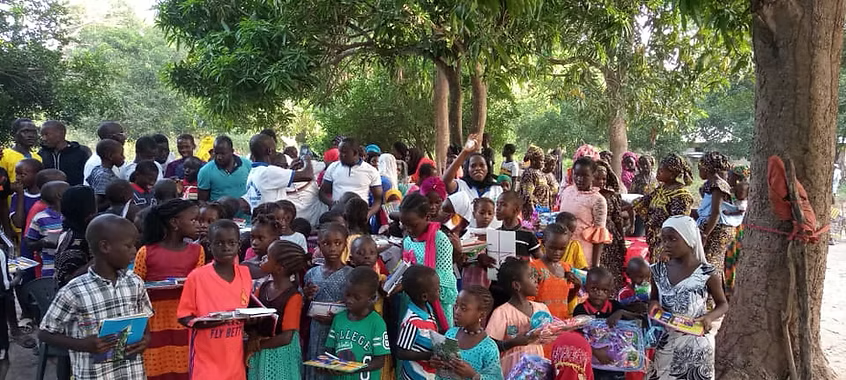 Crowd gathered outdoors under trees, some seated and standing, with colorful clothing and containers.