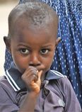 Young child in a gray shirt looking at the camera with a finger near their mouth outdoors.
