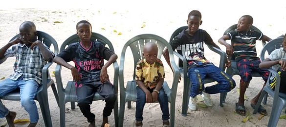 Six boys sitting on plastic chairs outdoors, posing with drinks in hand.