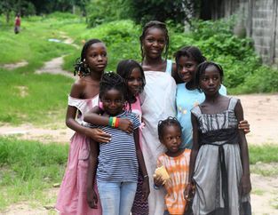 Seven children posing together on a grassy path, with trees and a wall in the background.