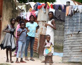 Five children stand in a narrow alley beside corrugated metal walls, with clothes hanging overhead.