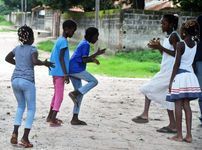 Children playing hopscotch outdoors on a dirt path, with a concrete wall and greenery in the background