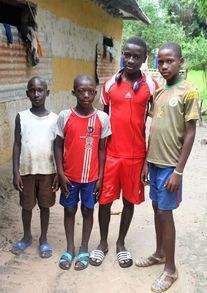 Four children stand outside a rustic building, wearing colorful casual clothes and sandals.