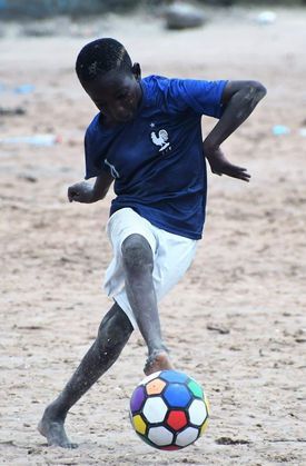 Boy in blue shirt and white shorts kicks a colorful soccer ball on a sandy field.