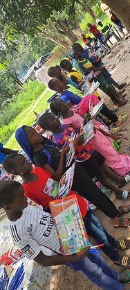People lined up outdoors holding books and supplies under trees, likely at a community distribution event.
