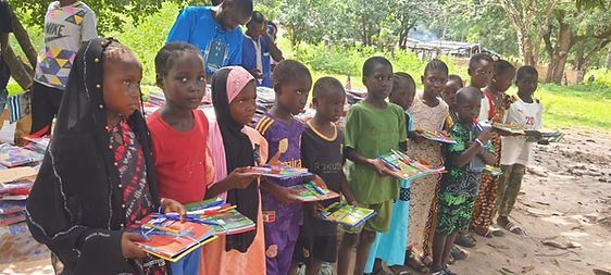 Children standing outdoors in a line, holding colorful schoolbooks and notebooks in a grassy area.