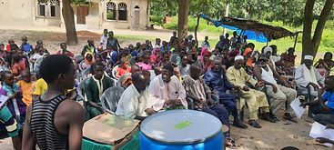 Crowd seated outdoors listening to a speaker, with blue drums in the foreground and a building behind.