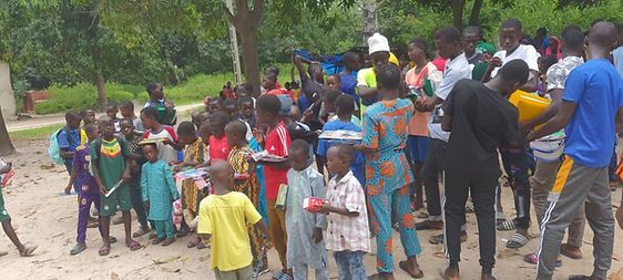 Crowd gathered outdoors near a dirt road, with people standing in colorful clothing and hats.