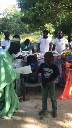 People gathered outdoors under trees around a table with items, one person in green in the foreground.