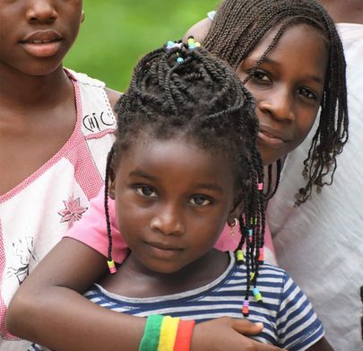Three children standing close together, with the front child wearing a striped shirt and beaded braids.
