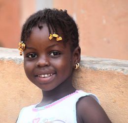 Smiling child with braided hair and yellow beads, standing by a peach-colored wall.