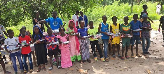 Group of children and adults standing outdoors in a line, holding a long yellow ribbon in a green setting