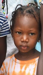 Young child with braided hair in an orange-and-white shirt, looking at the camera outdoors.