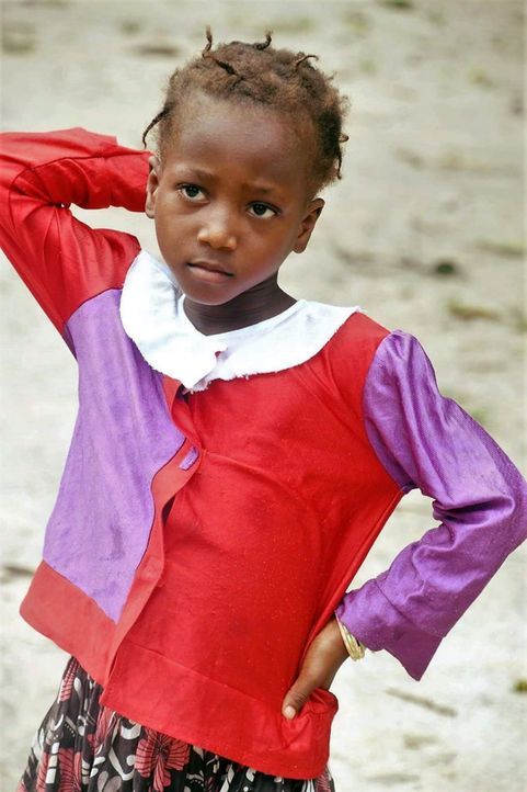 Child standing outdoors with hand on head, wearing a red, purple, and white shirt and patterned skirt.