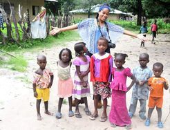 Woman with outstretched arms stands behind a group of children outdoors on a dirt path.
