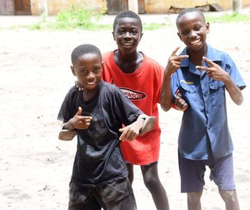 Three children smiling and posing outdoors, two making hand signs; one in red shirt, one in blue, one in black.