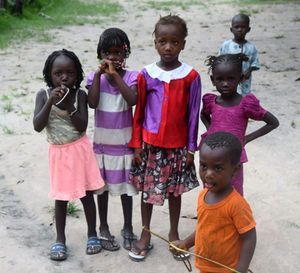 Five children standing outdoors on a dirt path, wearing colorful clothes and looking toward the camera.
