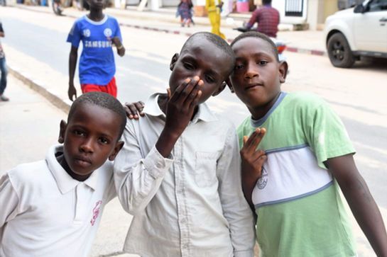 Three boys pose on a street, one making a peace sign, with cars and pedestrians in the background.