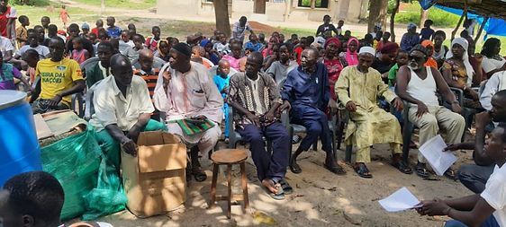 Crowd seated outdoors on plastic chairs during a community gathering, with papers scattered on the ground.