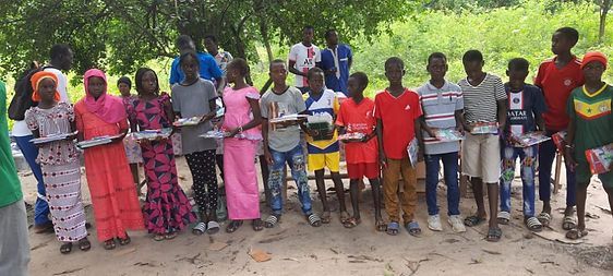 Group of people standing outdoors in a line, holding papers, with trees and sandy ground in the background.