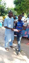 Two people standing outdoors, holding a document in a sunny street market area.