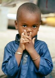 Child in a blue shirt holding hands near their face, looking directly at the camera.