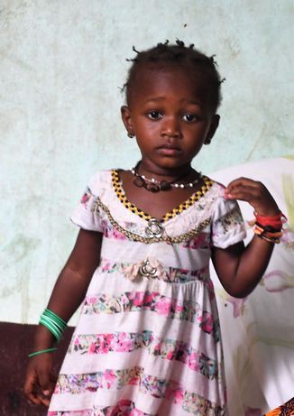 Young child in a pink floral dress standing against a plain wall, wearing bracelets and necklaces.