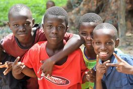 Four smiling children standing close together outdoors, one in a red shirt and another making a peace sign