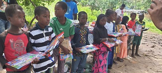Children holding colorful books stand in line outdoors under trees.