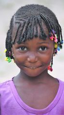 Smiling child with beaded braids wearing a purple shirt against a plain background