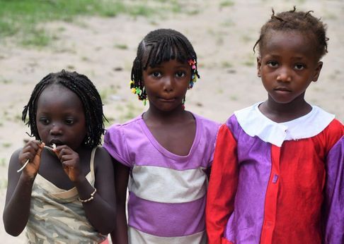 Three children standing outdoors, one eating a snack, with a blurred path in the background.