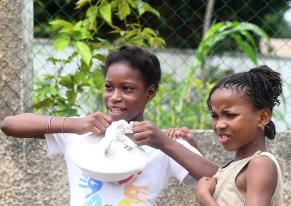 Two children outdoors, smiling and holding a white bowl or bucket together.