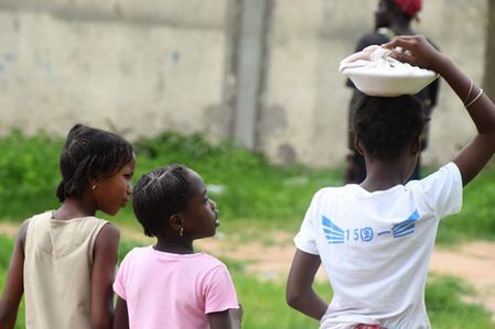 Three people walking outdoors, one balancing a white container on their head