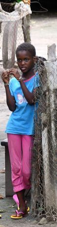 Child standing beside a tree, wearing a blue shirt and pink pants, holding a small white bag
