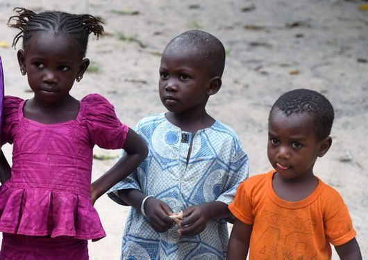 Three children standing outdoors, one in a pink dress, one in a blue shirt, and one in an orange shirt