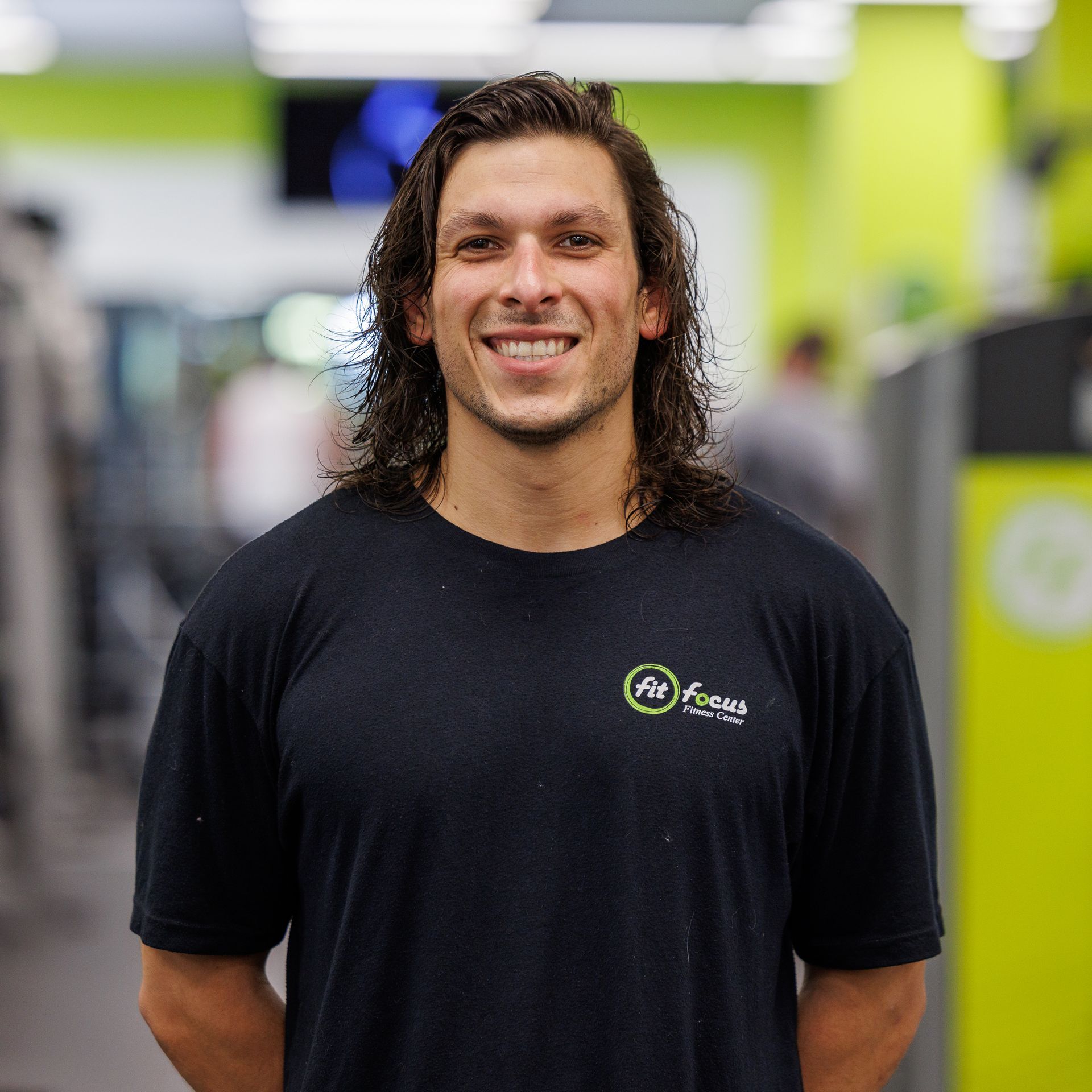 Man with long hair smiling in a gym, wearing a black t-shirt with a fitness logo.