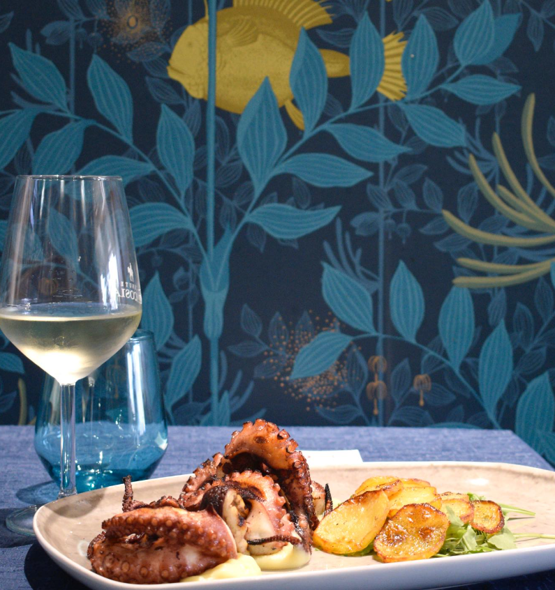 Piatto di polpo alla griglia con patate e bicchiere di vino su sfondo blu decorato.