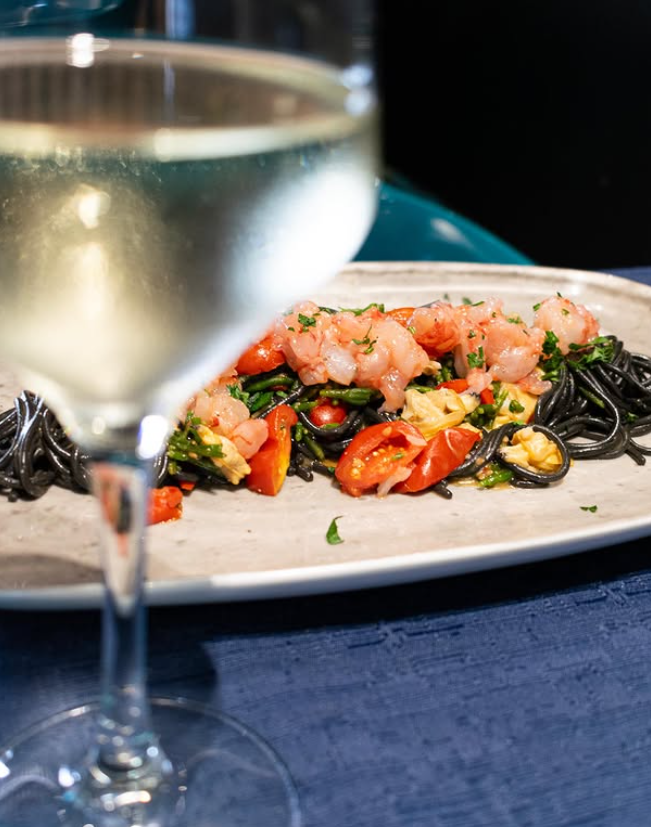 Piatto di spaghetti neri con gamberi e pomodori, accompagnato da un bicchiere di vino bianco.