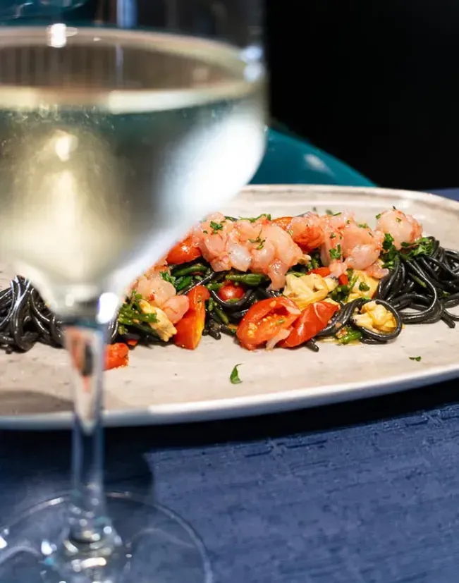 Piatto di pasta nera con gamberi, pomodori e prezzemolo, accompagnato da un bicchiere di vino bianco.