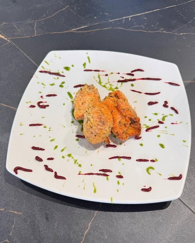 Cibo fritto su un piatto bianco, decorato con salse verdi e viola, posto su un bancone scuro.