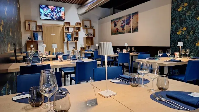 Interno del ristorante: tavoli apparecchiati con sedie blu, lampade e bicchieri. Le decorazioni includono quadri incorniciati e decorazioni murali.