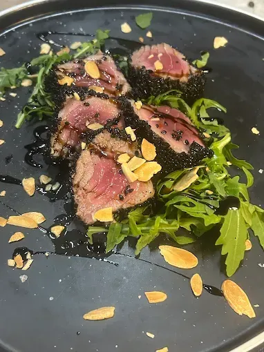 Tonno scottato con semi di sesamo, rucola, glassa balsamica e mandorle su un piatto nero.