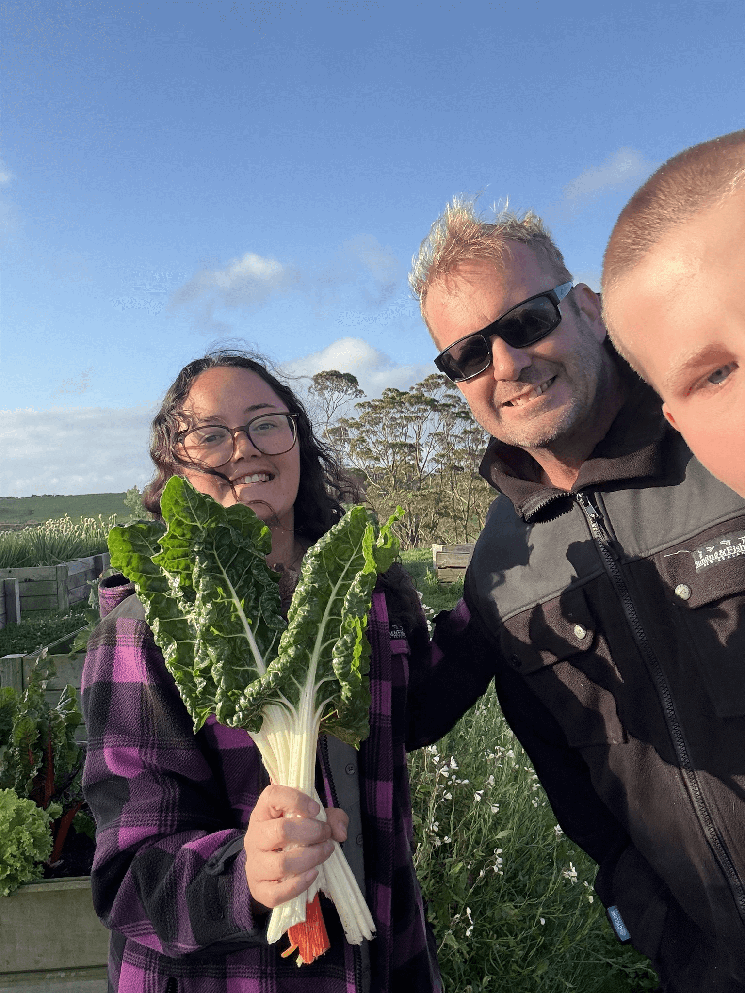 Group of three smiling people outdoors; one holding leafy green vegetables.