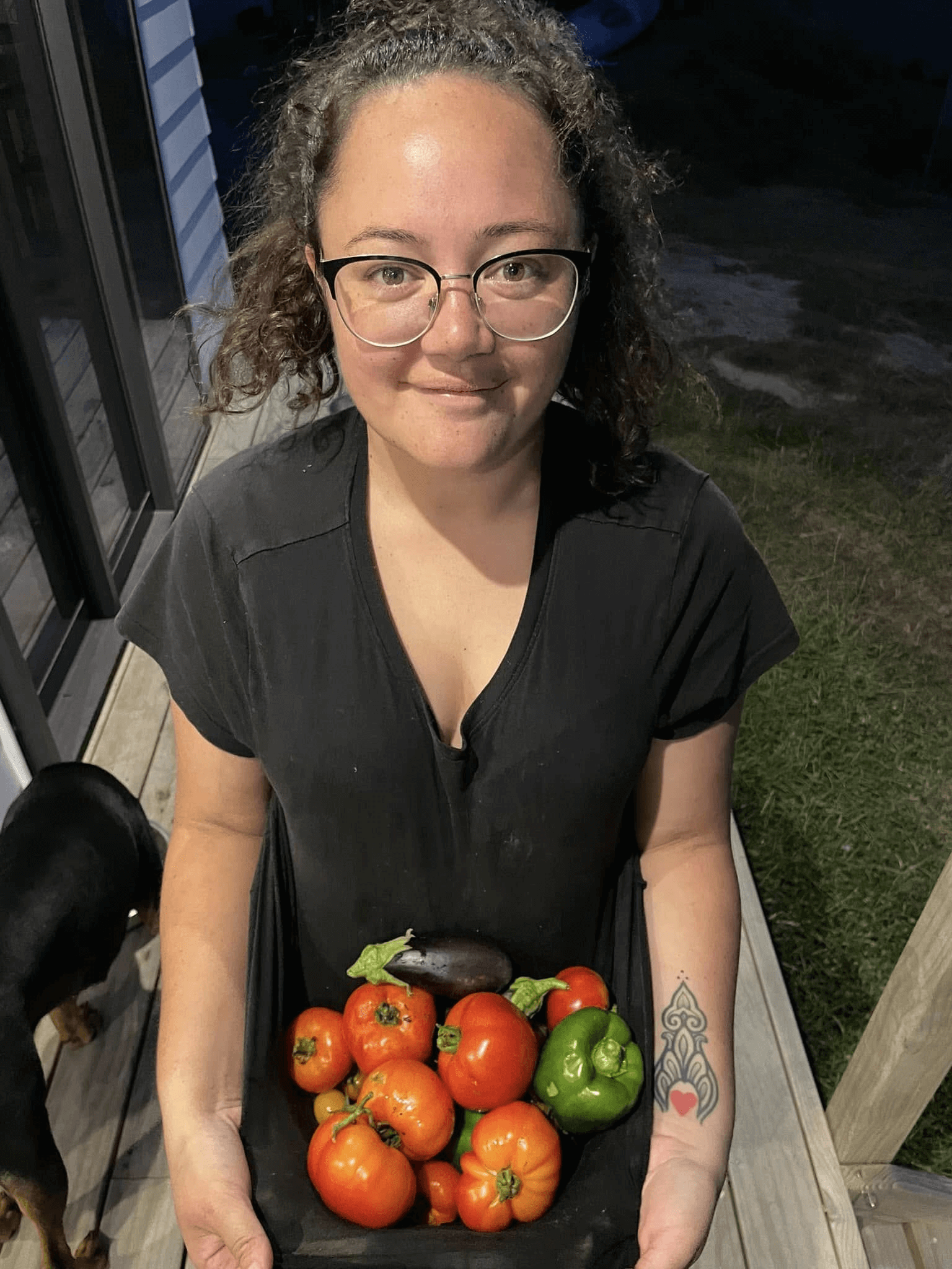 Woman holding a basket of fresh tomatoes, eggplant, and pepper. Outdoors, dog nearby.