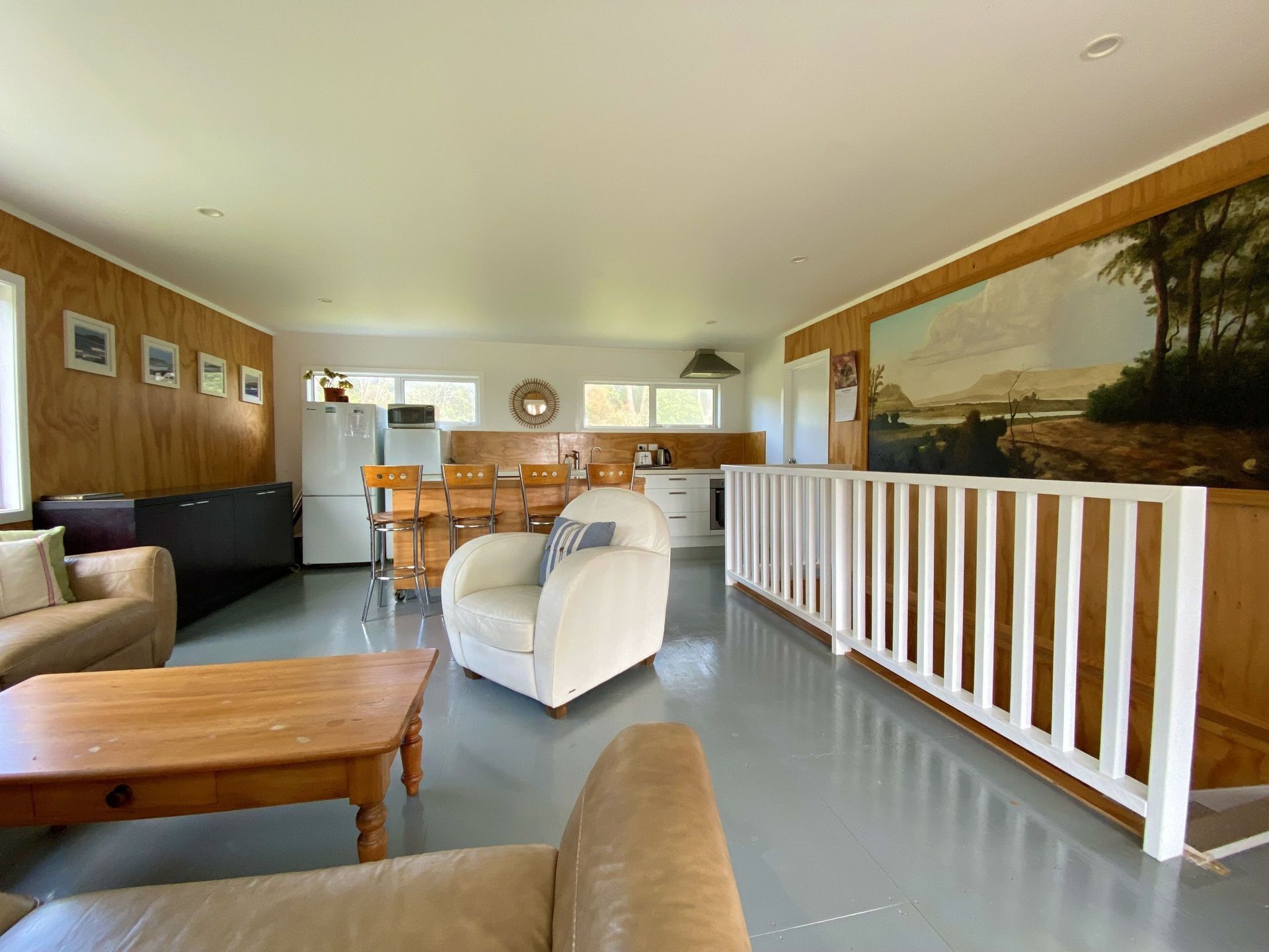 Open-plan living space with kitchen, seating area, and painting on the wall; gray floors and light-colored walls.