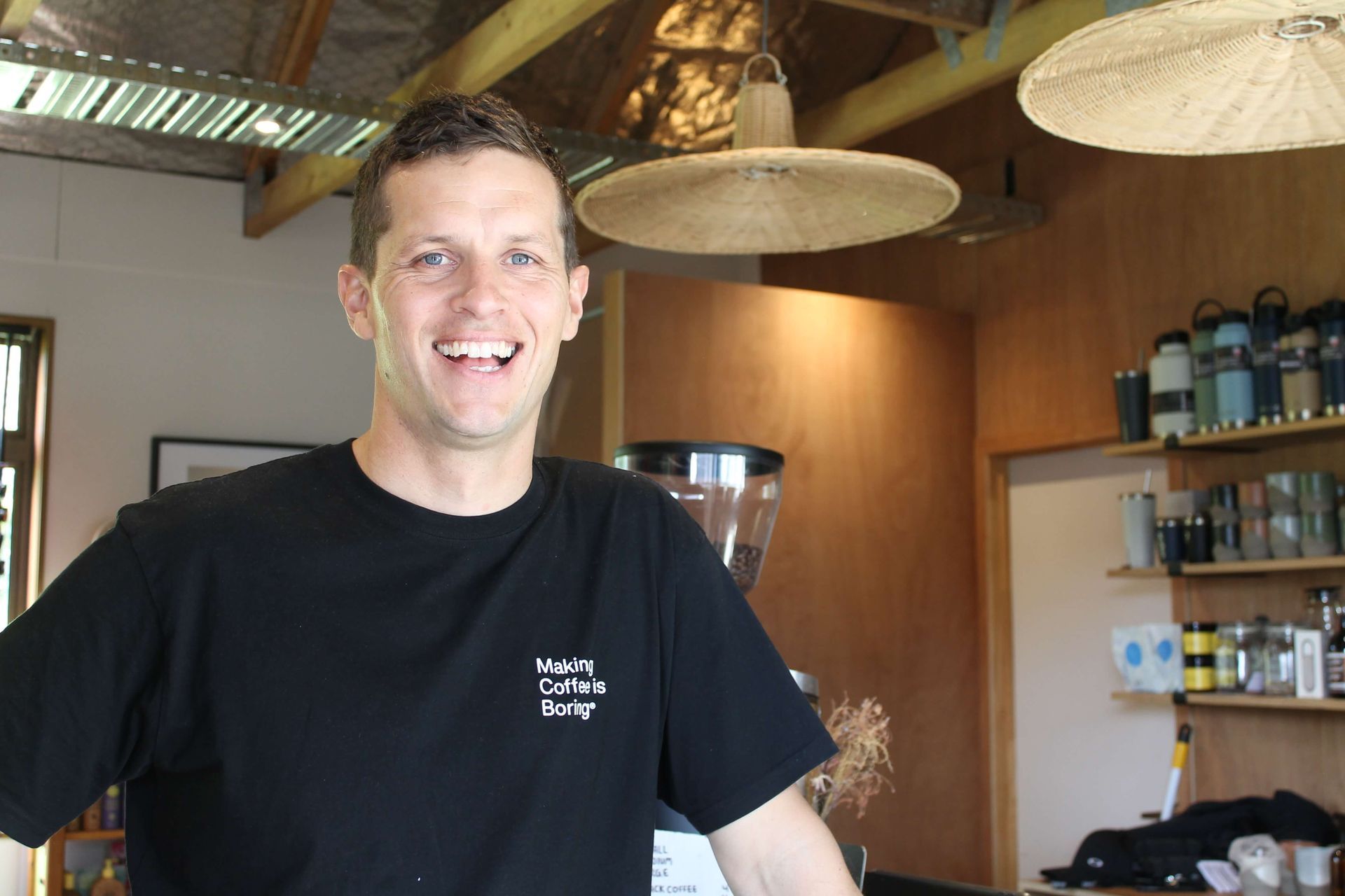 Man in black shirt smiling at a coffee shop. Wooden interior with hanging lamps, coffee machine, shelves.