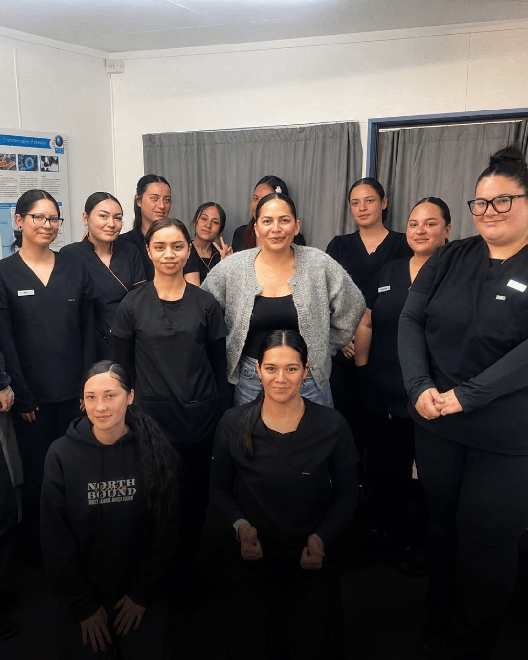 Group of people in black uniforms posing indoors in a small room with curtains.